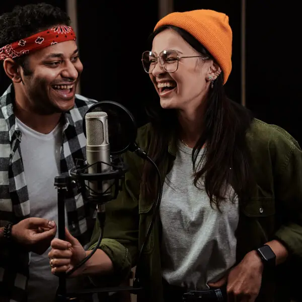 Portrait of cheerful young man and woman, duet singing into a condenser microphone while recording a song in a professional studio. Selective focus. Horizontal shot